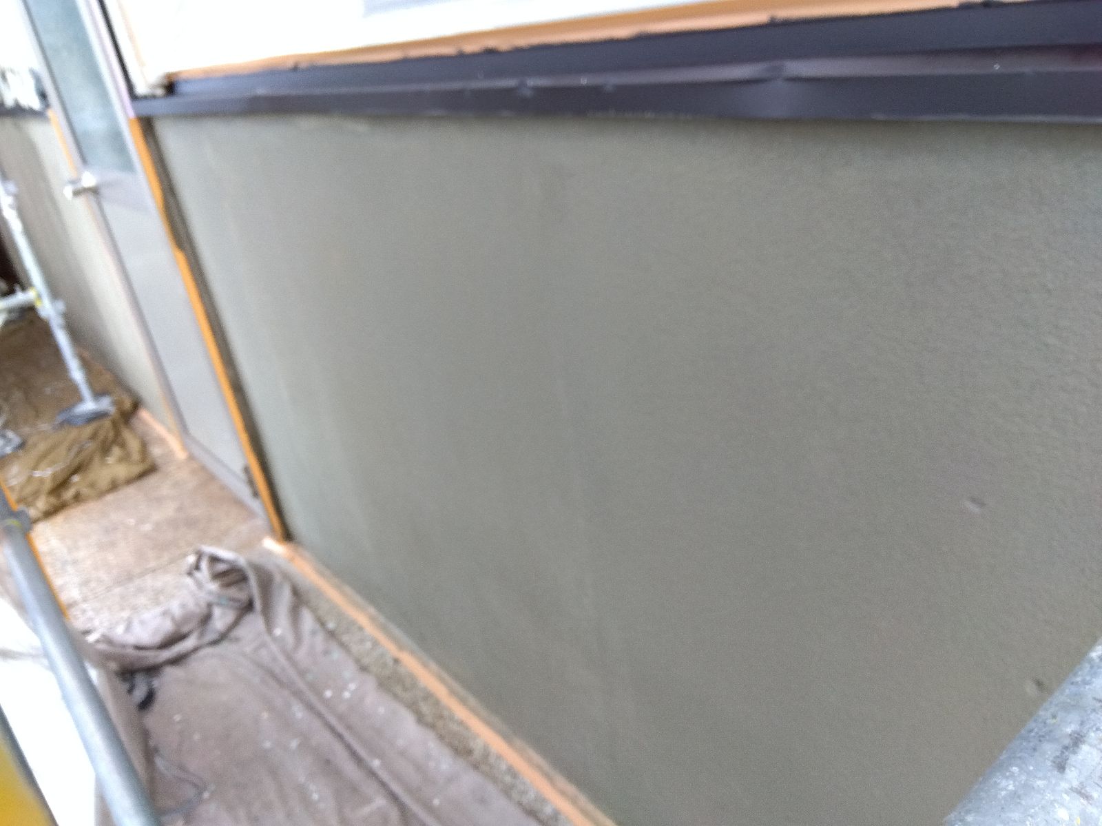 0b68a084-925a-4b1a-bf3d-113f04d9b5b0 | 岐阜県大垣市 塗りかえ工房 Close-up of a grey plastered exterior wall under construction, with orange trim tape along the edges and a metal door frame on the left; construction debris on the ground.