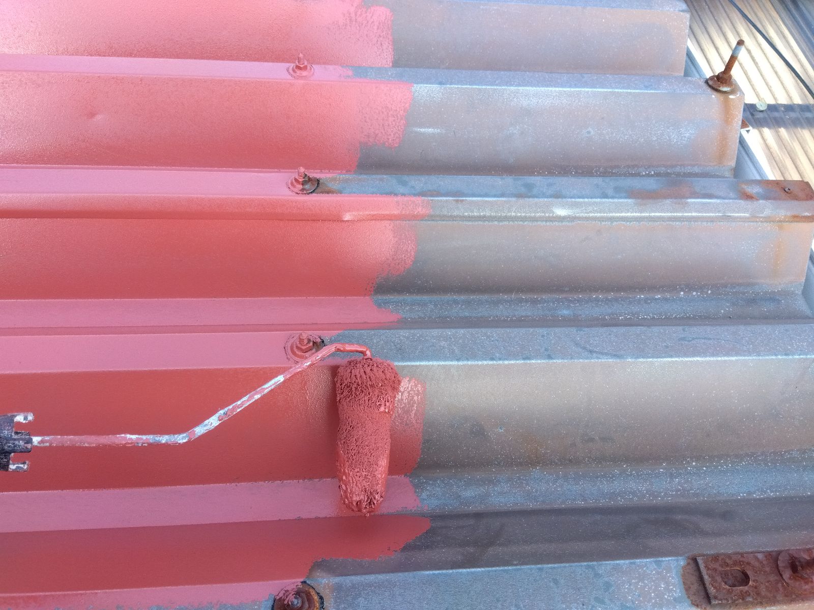 1466929a-2c48-4fce-9672-a84b0c624385 | 岐阜県大垣市 塗りかえ工房 Metal corrugated stairs being painted red using a roller, with partially covered red paint on the left and blue-gray metal on the right; bolts show rusty edges.
