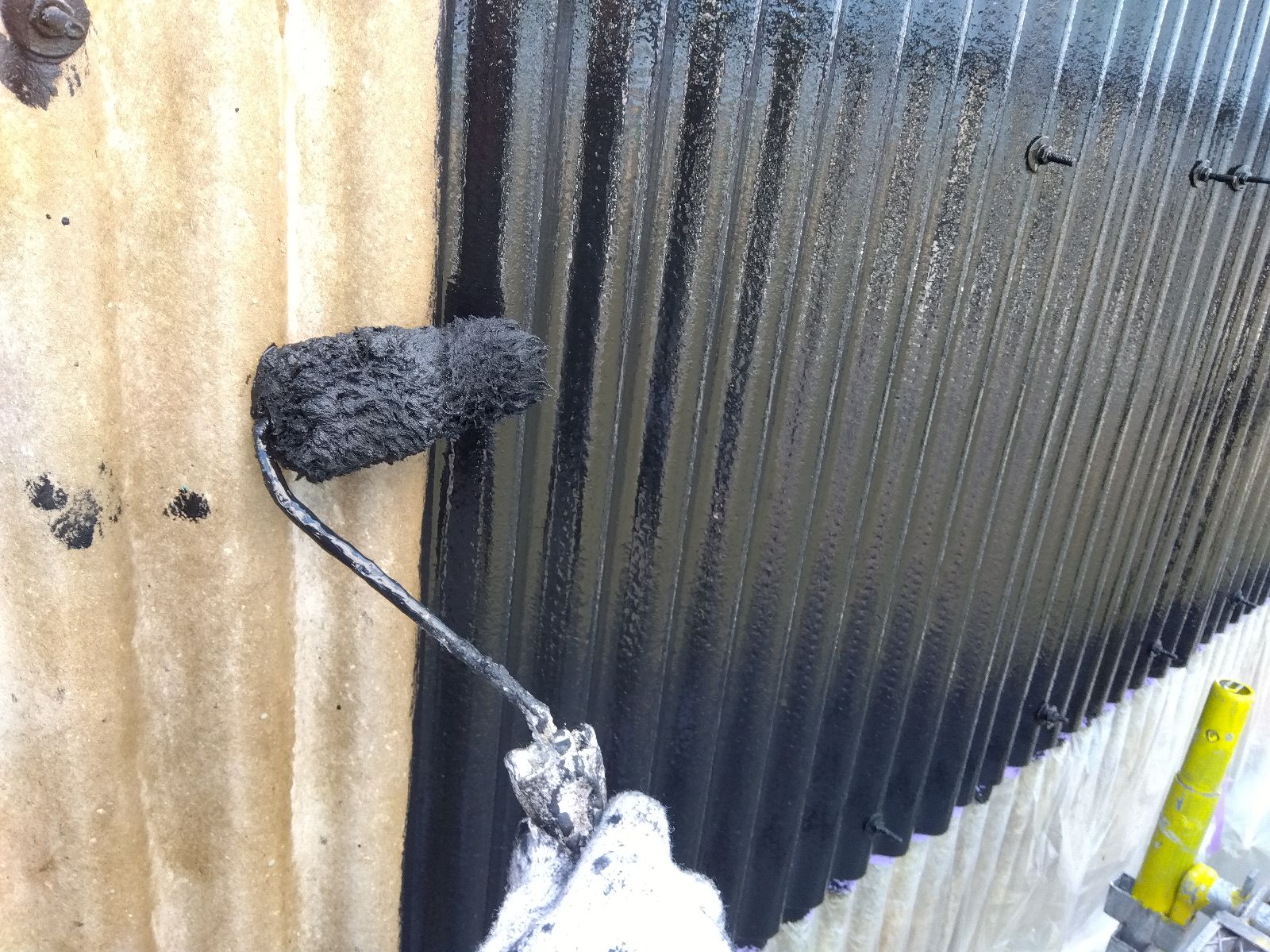 76e31b3b-9e9a-4b40-b3a4-d0d83b3066fb | 岐阜県大垣市 塗りかえ工房 Black paint is being rolled onto a corrugated metal wall with a paint roller; wet paint visible on the surface and left side shows a brown wall edge.