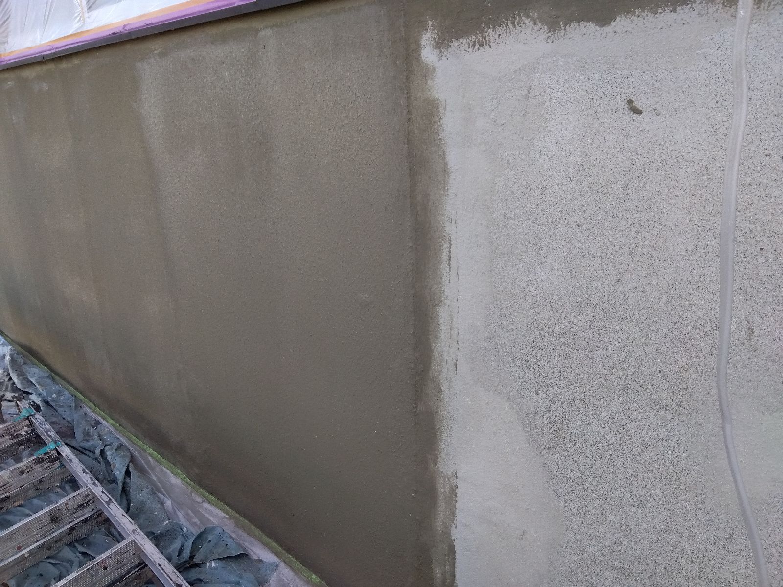 e4368e85-10c9-47be-a945-e1d81545a441 | 岐阜県大垣市 塗りかえ工房 Concrete wall with fresh brown plaster on left and lighter gray cement on right; construction tarp and wooden ladder at bottom left edge, top taped with purple strip.