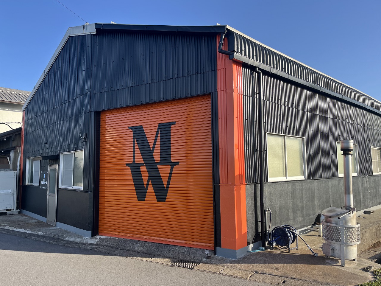 20250322マツバラワーク1-2 1280x960 | 岐阜県大垣市 塗りかえ工房 Industrial warehouse with a bright orange roll-up door featuring the letters MW against a dark corrugated metal exterior.