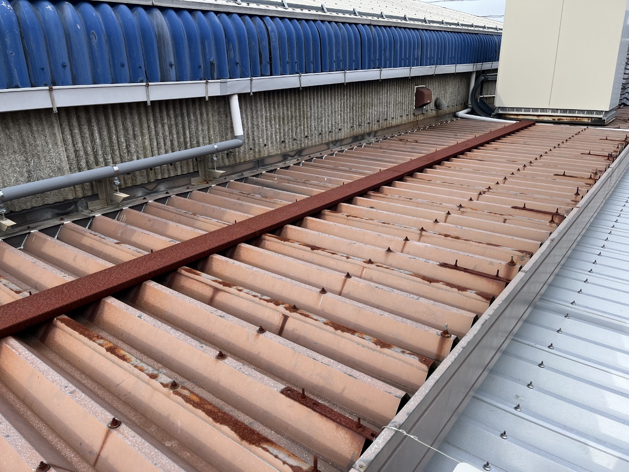 20250322マツバラワーク4-1 1280x960 | 岐阜県大垣市 塗りかえ工房 Rooftop with brown, rusty corrugated metal panels, a long red steel beam, blue curved roof vents along the wall, and an outdoor HVAC unit in the background.