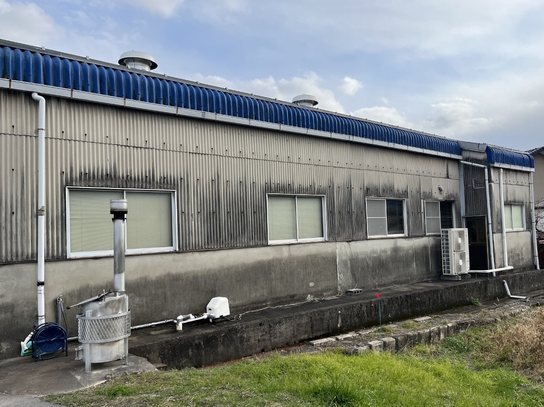 20250322マツバラワーク3-1 1280x960 | 岐阜県大垣市 塗りかえ工房 Industrial building exterior with a blue corrugated roof, gray metal walls, and a row of windows along a concrete base.