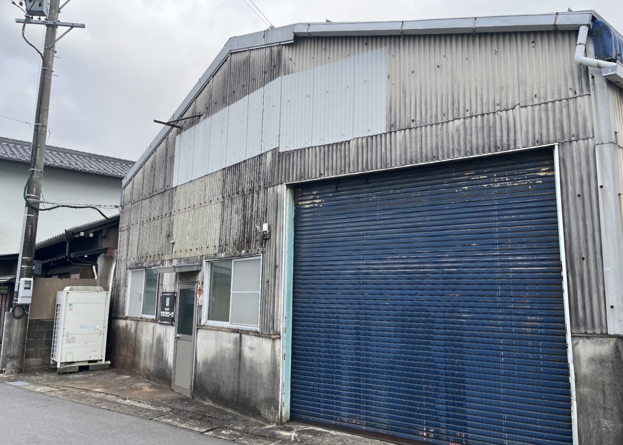 20250322マツバラワーク2-1 1280x960 | 岐阜県大垣市 塗りかえ工房 Industrial corrugated-metal warehouse with a large blue rolling shutter door and small side door, weathered exterior and utility pole nearby