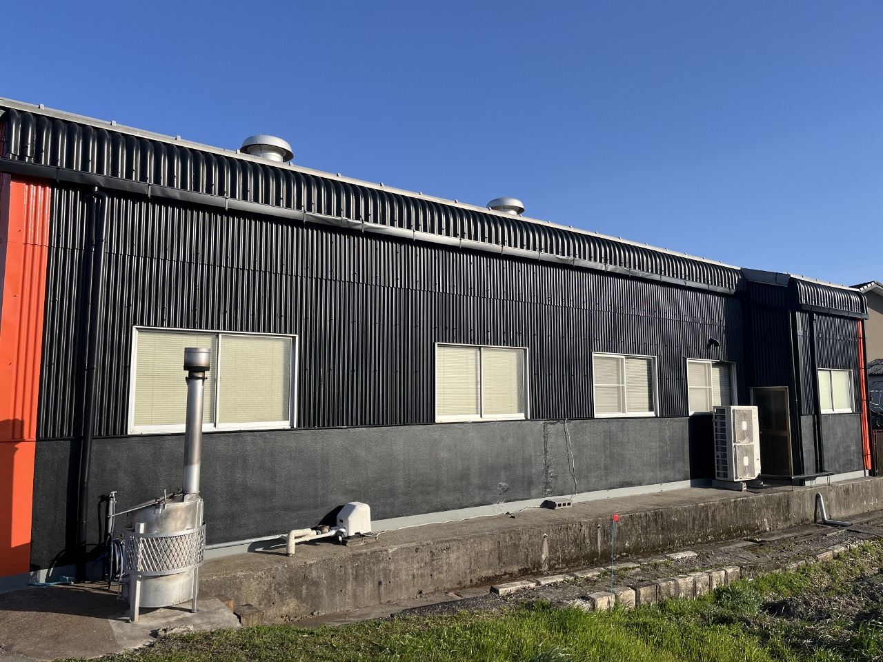 20250322マツバラワーク3-2 1280x960 | 岐阜県大垣市 塗りかえ工房 Industrial gray metal building with corrugated panels, several small windows, and an air conditioner unit along a concrete platform against a clear blue sky.