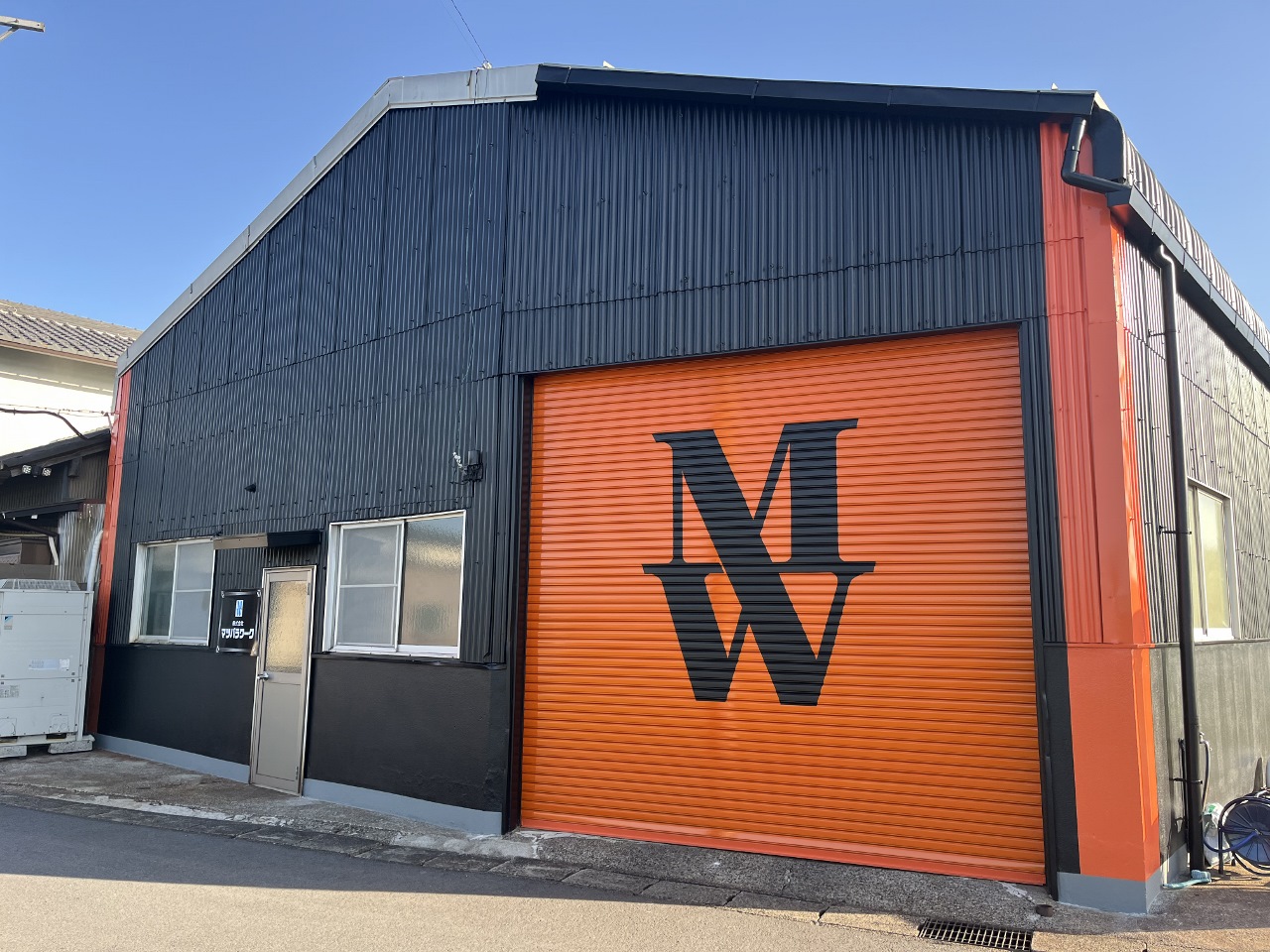 Industrial building with dark blue corrugated metal siding and a large orange roller door featuring a bold black 'MW' logo.
